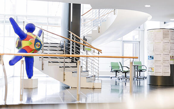 Das helle Foyer des Ärztehauses. Man sieht den Treppenaufgang, daneben steht eine große bunte, runde Figur, die ihre Arme und Beine ausstreckt. Auf der rechten Seite des Bildes befindet sich eine Säule mit Werbeplakaten.