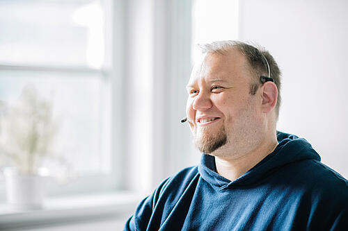 Ein Mann mit kurzen Haaren und einem dunklen Pullover sitzt an einem Schreibtisch mit einem Headset auf. Er telefoniert und lächelt dabei.