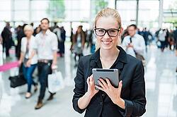 Das Bild zeigt eine Frau auf einer Messe. Sie hält ein Tablet in der Hand, schaut in die Kamera, Im Hintergrund gehen viele Menschen.