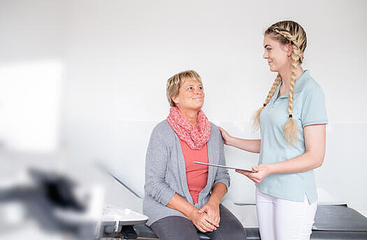 Eine junge MFA beruhigt eine Patientin, die auf einer Liege sitzt, indem sie ihre Hand auf die Schulter legt