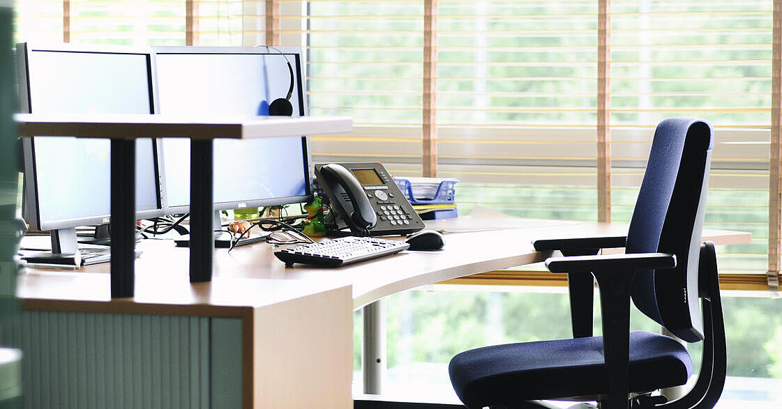 Ein leeres helles Büro, in dem man einen Schreibtisch und einen Schreibtischstuhl sieht. Im Vordergrund steht ein Aktenschrank.