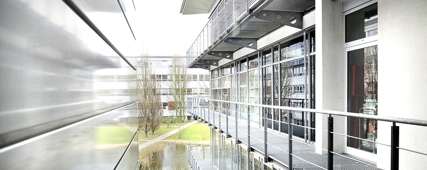 Blick zwischen zwei Gebäudehälften. Auf der rechten Seite sieht man eine Reihe von Fenstertüren, davor ein Gitterrost mit Geländer. Auf der rechten Seite eine verkleidete Gebäudewand.