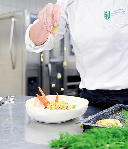 Eine gefüllt Salatbowl steht auf einem Tisch, daneben ein Bund Petersilie. Hinter dem Tisch sieht man eine Frau, die eine weiße Kochjacke mit dem Logo der Ärztekammer trägt. Man sieht noch ihre Hand, die kleine Salatstücke in die Bowl herunterrieseln lässt. DAs Bild endet über der Brust der Frau, der Kopf ist nicht mehr zu sehen.