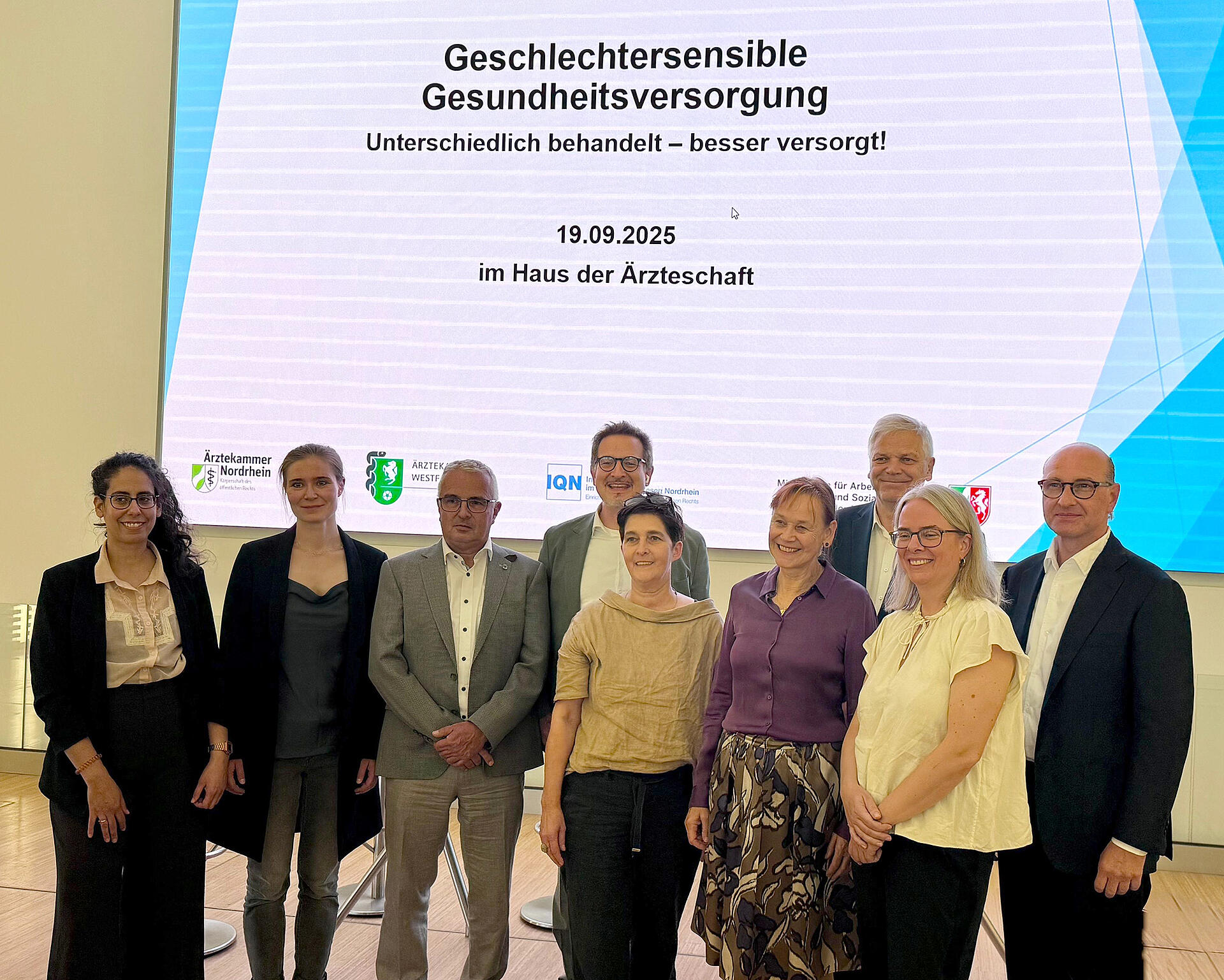Gruppe mit Männern und Frauen stehen vor einer Wand, auf der der Titel der Veranstaltung "Geschlechtersensible Gesundheitsversorgung" projiziert wird.