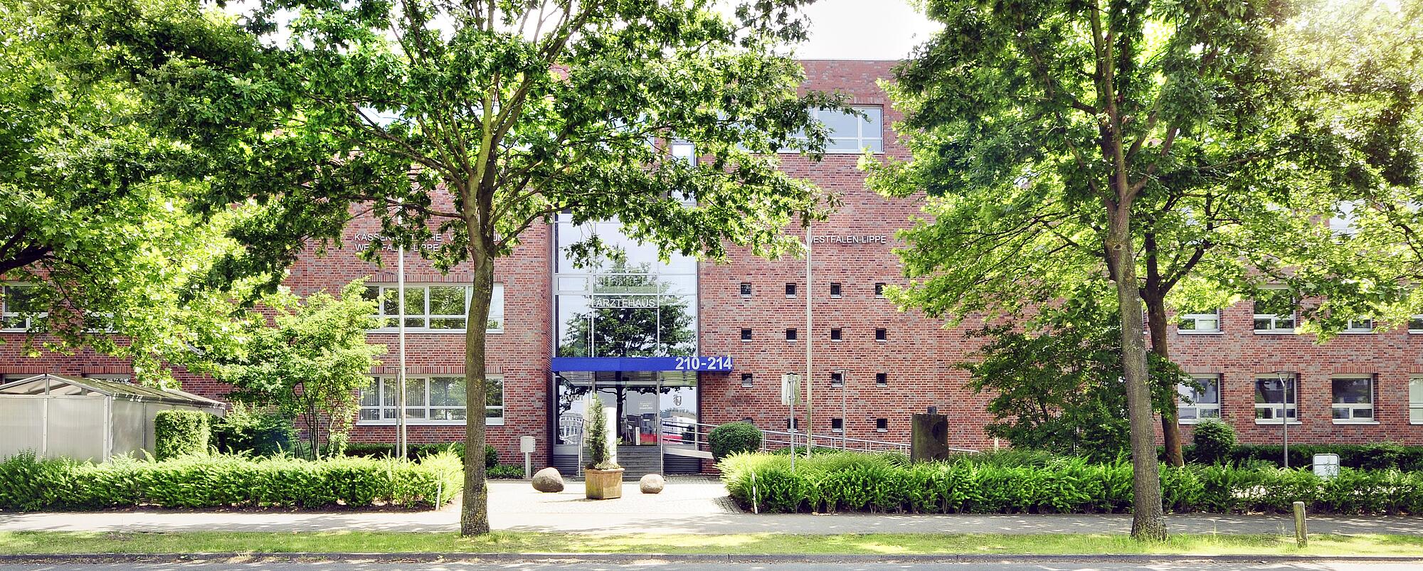 Der Haupteingang des Ärztehauses in Münster, Blick von der Straße aus. Man sieht viele Baäume, dahinter das Gebäude, groß über dem Eingang die Hausnummer auf blauem Grund "210 - 214".