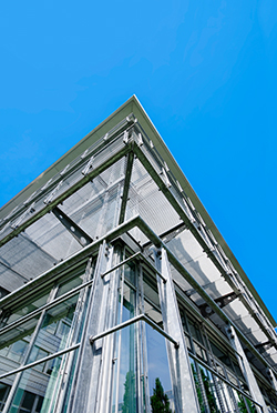 Die Ecke eines Gebäudes von unten fotografiert. Das Gebäude ist von einer Stahlkonstruktion umfasst, man sieht außerdem große Fenster und einen sehr blauen Himmel.