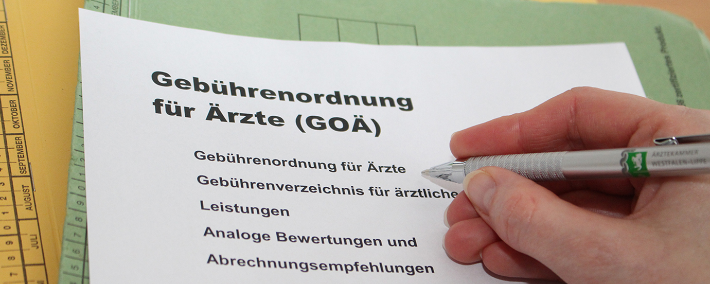 Ein Zettel mit dem Text "Gebührenordnung für Ärzte" liegt auf zwei Akten. Man sieht außerdem eine Hand, die einen Kugelschreiber mit dem Logo der Ärztekammer Westfalen-Lippe hält. 