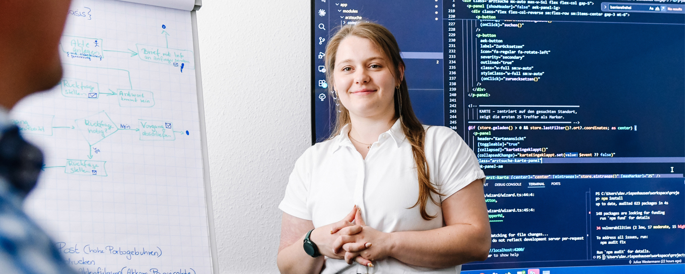 Lächelnde Frau in weißer Bluse steht zwischen einem Flipchart mit handgezeichneten Ablaufdiagrammen und einem großen Bildschirm mit Programmcode.