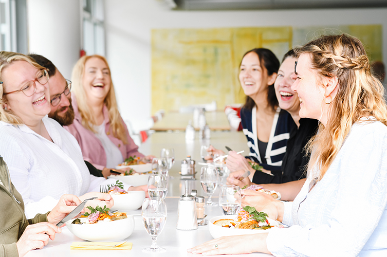 Sechs Menschen sitzen zusammen an einem gedeckten Tisch. Sie unterhalten sich und lachen. Auf dem Tisch stehen Salatbowls und Gläser.