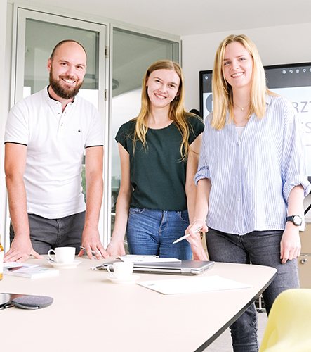 Ein junger Mann und zwei junge Frauen stehen in einem Besprechungsraum, vor ihnen ein Tisch mit Akten, Laptop und Schreibmaterial. Sie schauen in die Kamera und sehen fröhlich aus.