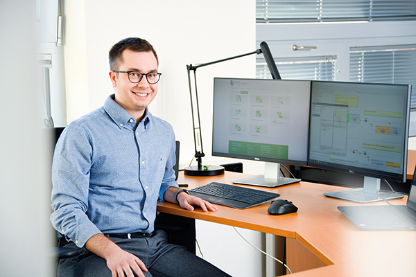 Ein junger Mann sitzt an seinem Schreibtisch, er lächelt in die Kamera. Auf dem Schreibtisch sieht man die Computermaus und -tastatur und zwei Bildschirme.