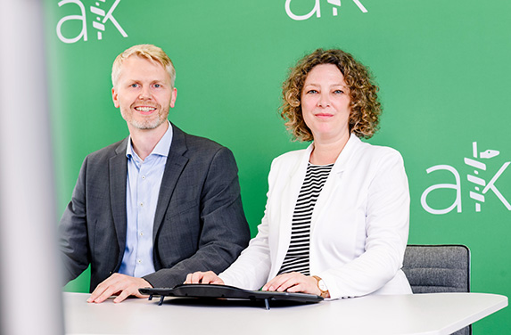 Ein Mann mit kurzen blonden Haaren und Jacket sitzt an einem weißen Schreibtisch. Daneben sitzt eine Frau mit lockigen Haaren und weißem Blazer. Beide lächeln in die Kamera. Im Hintergrund ist eine Blue Screen Wand zu sehen.