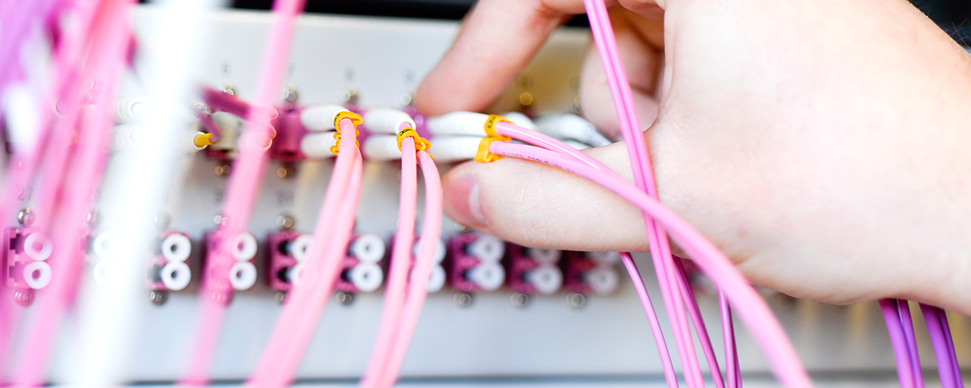 Der Serverraum. Zu sehen ist eine Hand, die ein rosa Kabel in eine Buchse steckt, daneben sieht man noch mehr Kabel.