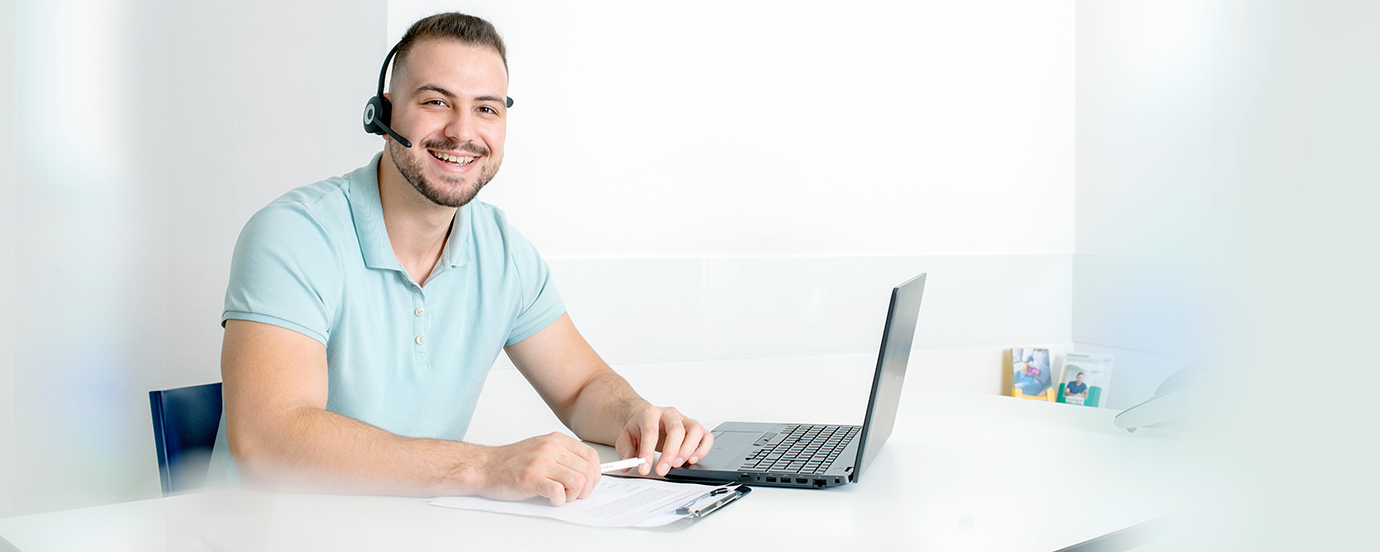 Medizinischer Fachangestellter mit Headset sitzt an einem Tisch. Vor ihm steht ein Laptop und in seiner Hand hält er einen Kugelschreiber. Er schaut in die Kamera.