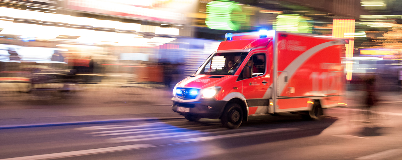 Ein Rettungswagen mit Blaulicht fährt durch eine belebte Straße an Einkaufsläden vorbei.