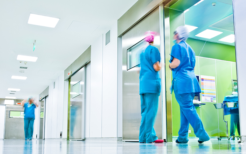 Krankenhaus, OP-Bereich. Im Vordergrund unterhalten sich zwei Frauen in OP-Kleidung, weiter hinten geht ein Mann in OP-Kleidung den  Gang hinunter. Ethikforum 2024. 